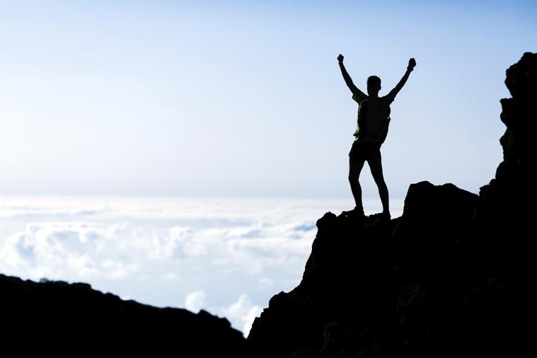 Man stands on top of mountain cliff raising hands in triumph