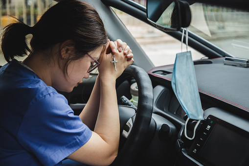 Female brunette nurse in the car praying with mask hanging from rearview mirror