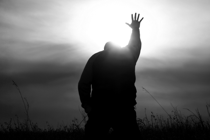 Black and white image of a man on his knees with one arm and hand raised up towards the sun